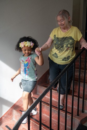 A senior woman and a girl aged about six are walking down a staircase holding hands. They are dressed in summer clothes and sunshine comes through a window