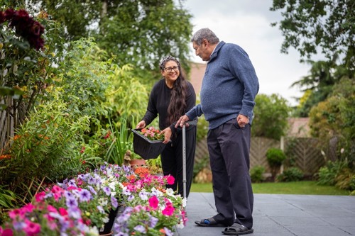 A man using a walking stick is talking to a woman who holds a tub of flowers. They are outside enjoying the flowers and plants in a beautiful garden border