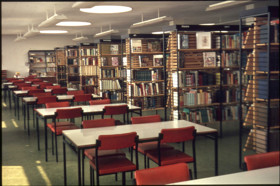 old photo of crosby library second floor with the left side having a line of tables and chairs and the right side having a line of metal bookshelves