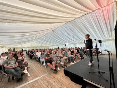 A shot of a crowd of people looking towards a stage. On the stage, and at the forefront of the picture is a man with a microphone.