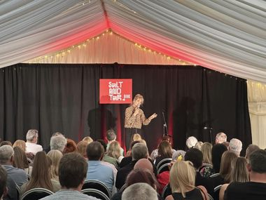 A picture taken from the back of a room. In front of the camera is a crowd of people, watching someone perform on stage.