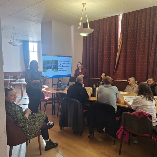 Photograph from the January 2026 Coproduction Meeting with a screen showing a presentation and people sat around the table