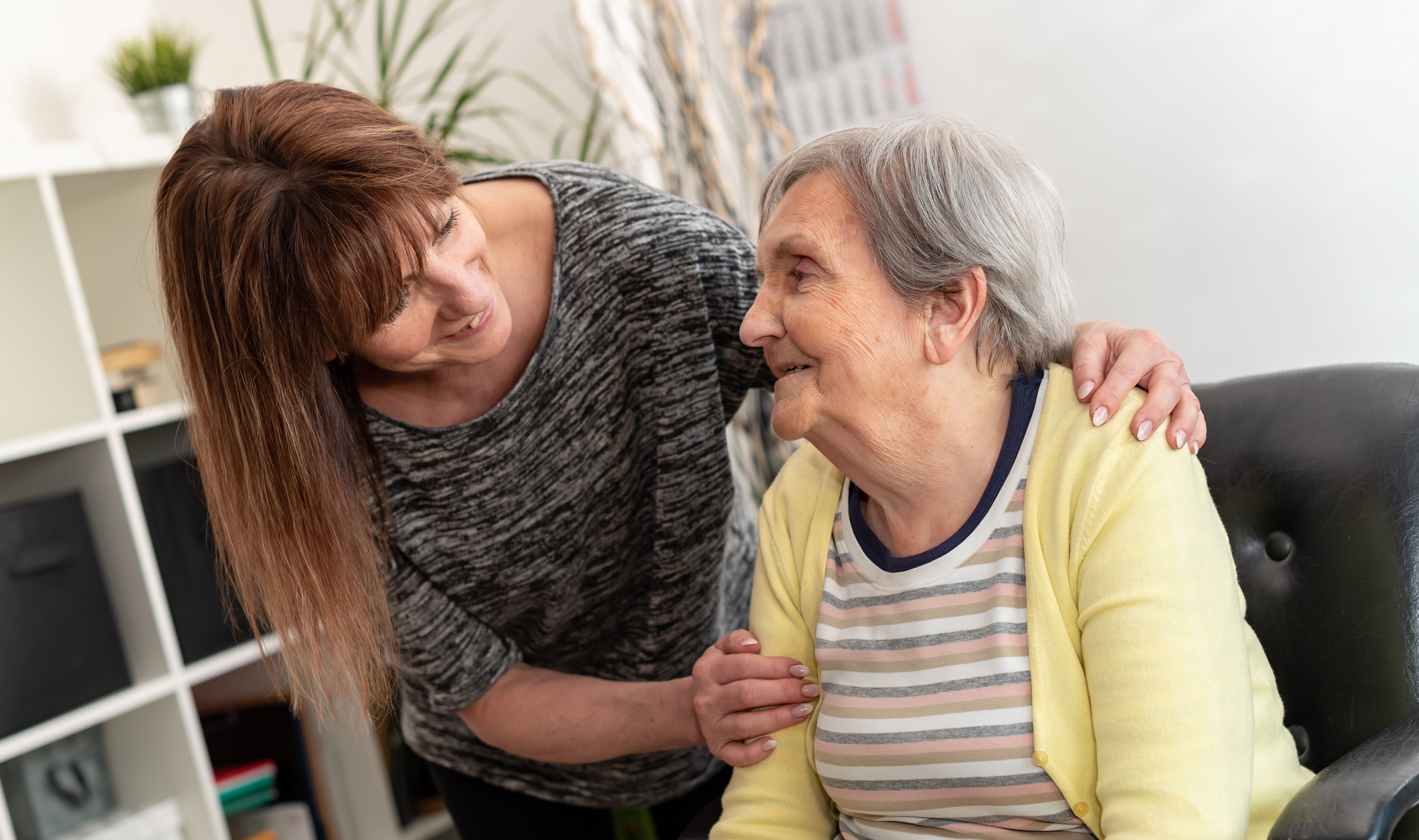 Adult gently supporting an older person while sitting together indoors in a home setting.