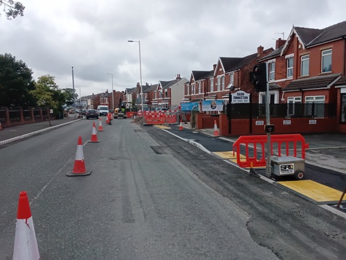 A photo of a road, half coned off. The pavement has freshly lain tarmac. There are some barriers in place for temporary crossing lights.