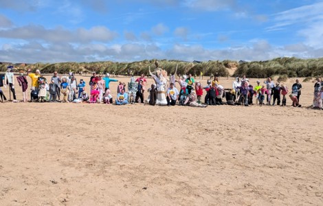 A photo on a beach of statues made of recycled wood and fabric alongside people.