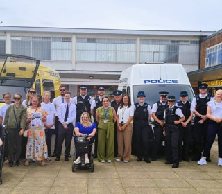 A large group of community members and police officers pose for a group photo in an outdoor shopping precinct. The group stands in front of two police vehicles, including a yellow police van on the left and a white police van on the right. Several adults are accompanied by young children, including one child seated in a pushchair and another partially visible beside it. One person is using a mobility scooter at the front of the group. 