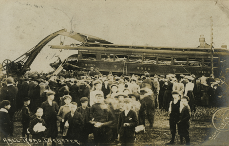 Sepia photo of crowd stood in front of wrecked train carriage