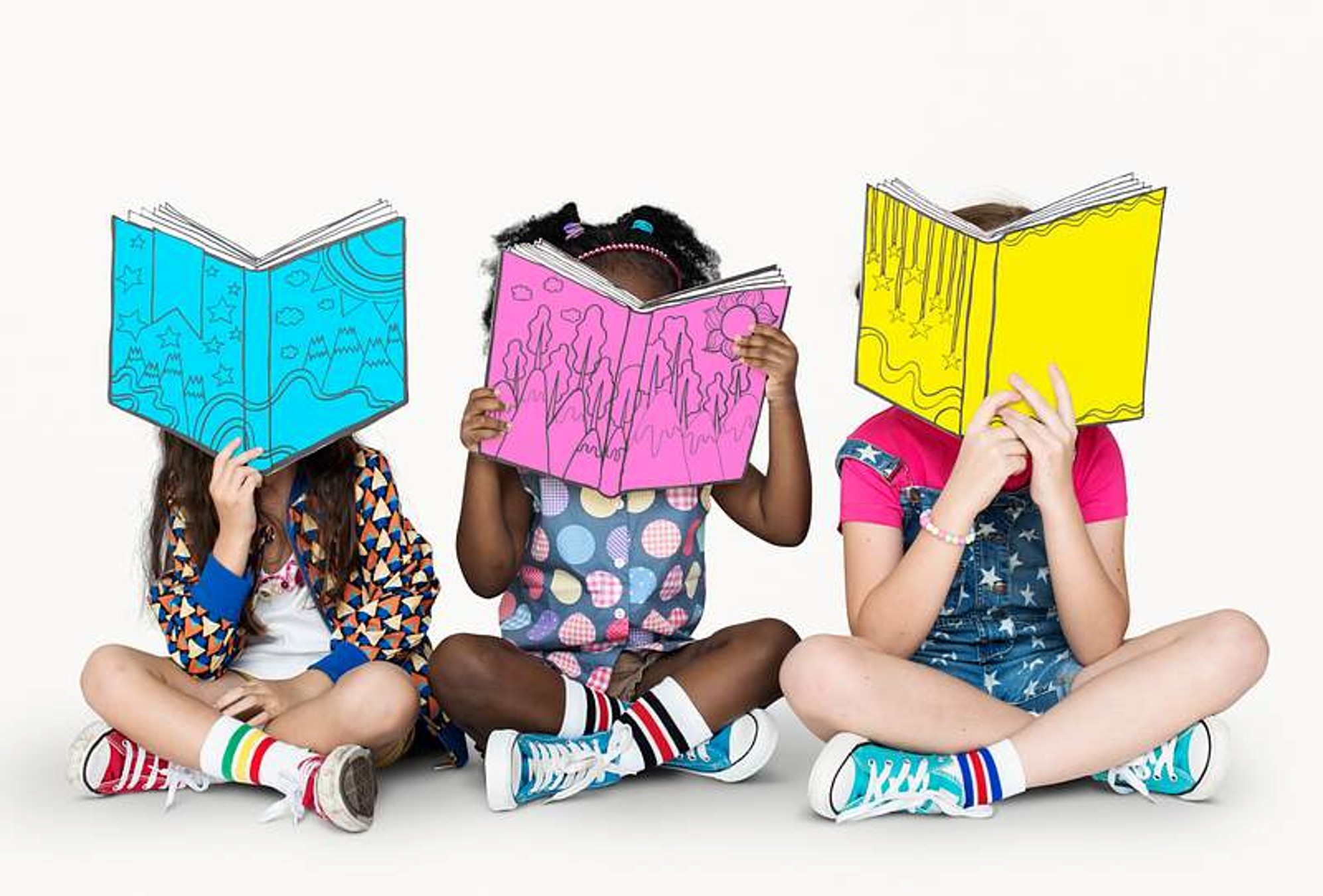 Three children sit cross-legged, each holding an open, colorful book in front of their faces. The books are blue, pink, and yellow, with simple line drawings on their pages.